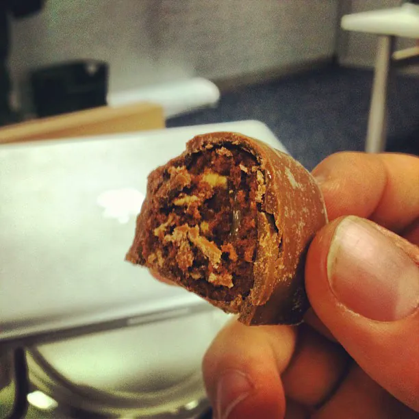 A hand holding a bitten chocolate pastry in front of a laptop