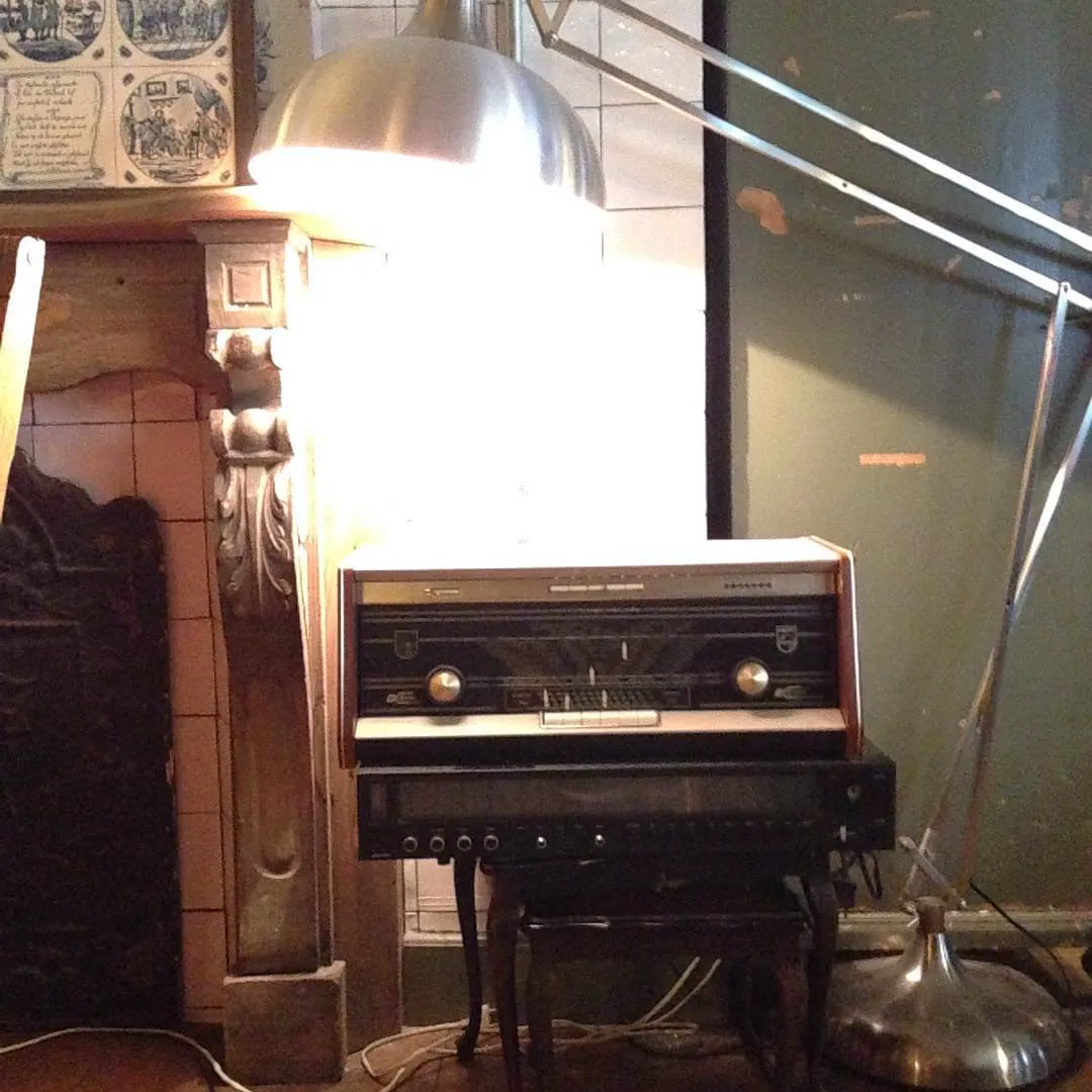 An old valve radio on a stand beside a carved fireplace and a lamp