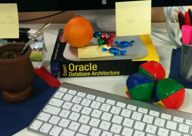 A desk with a keyboard, an orange, sweets, juggling balls, sticky notes and an Oracle database book